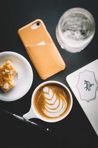cup of coffee selective focus photo of cup of cappuccino beside smartphone, saucer, and glass flat white